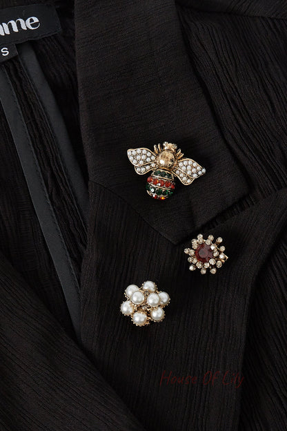 Three brooches pinned on a black blouse lapel: pearl-rhinestone bee with red and green accents, a clear red rhinestone flower, and a pearl cluster flower showcasing playful elegance