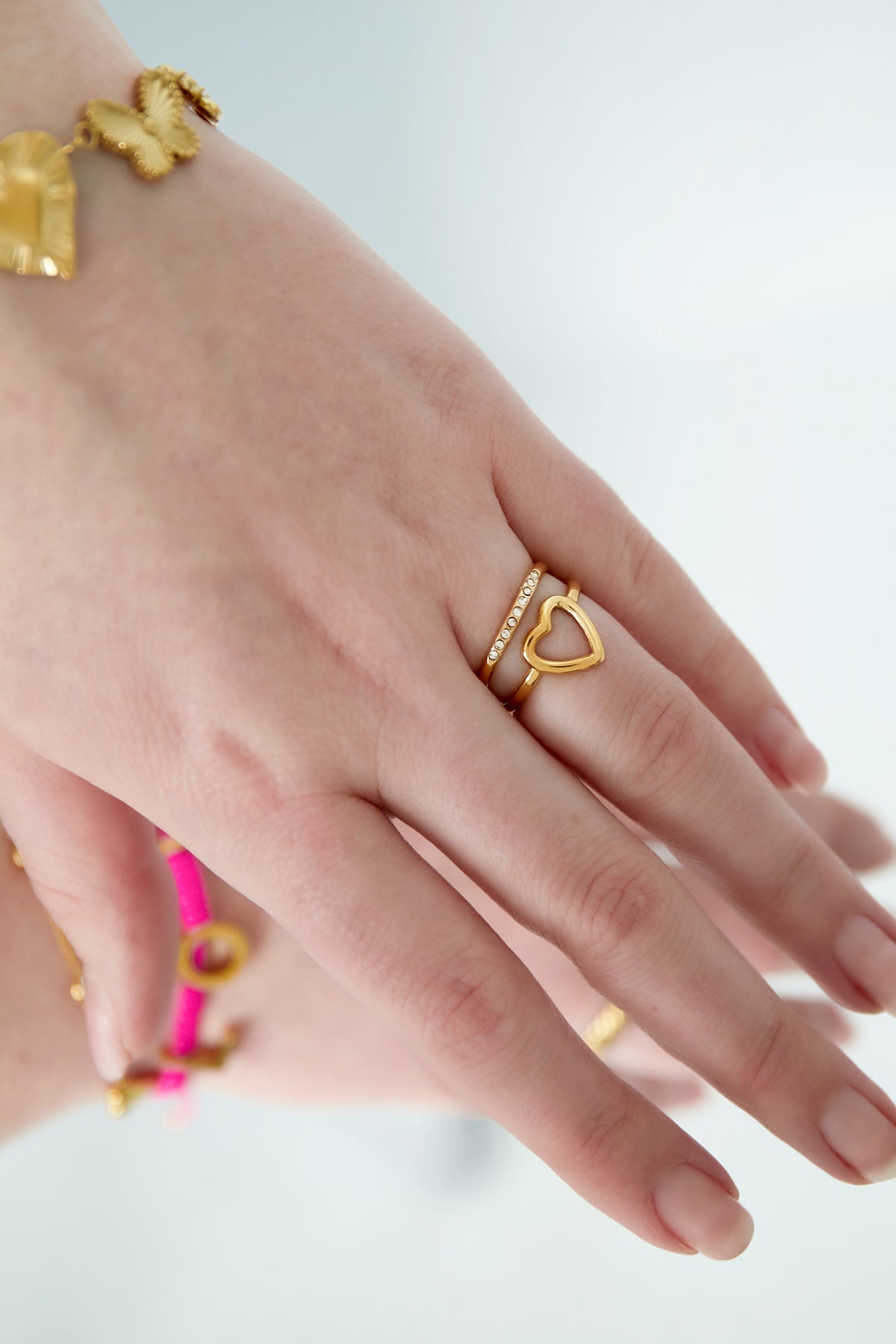 Side angle view of a woman's hand wearing the gold heart and rhinestone accent rings, accessorized with butterfly and heart charm bracelets