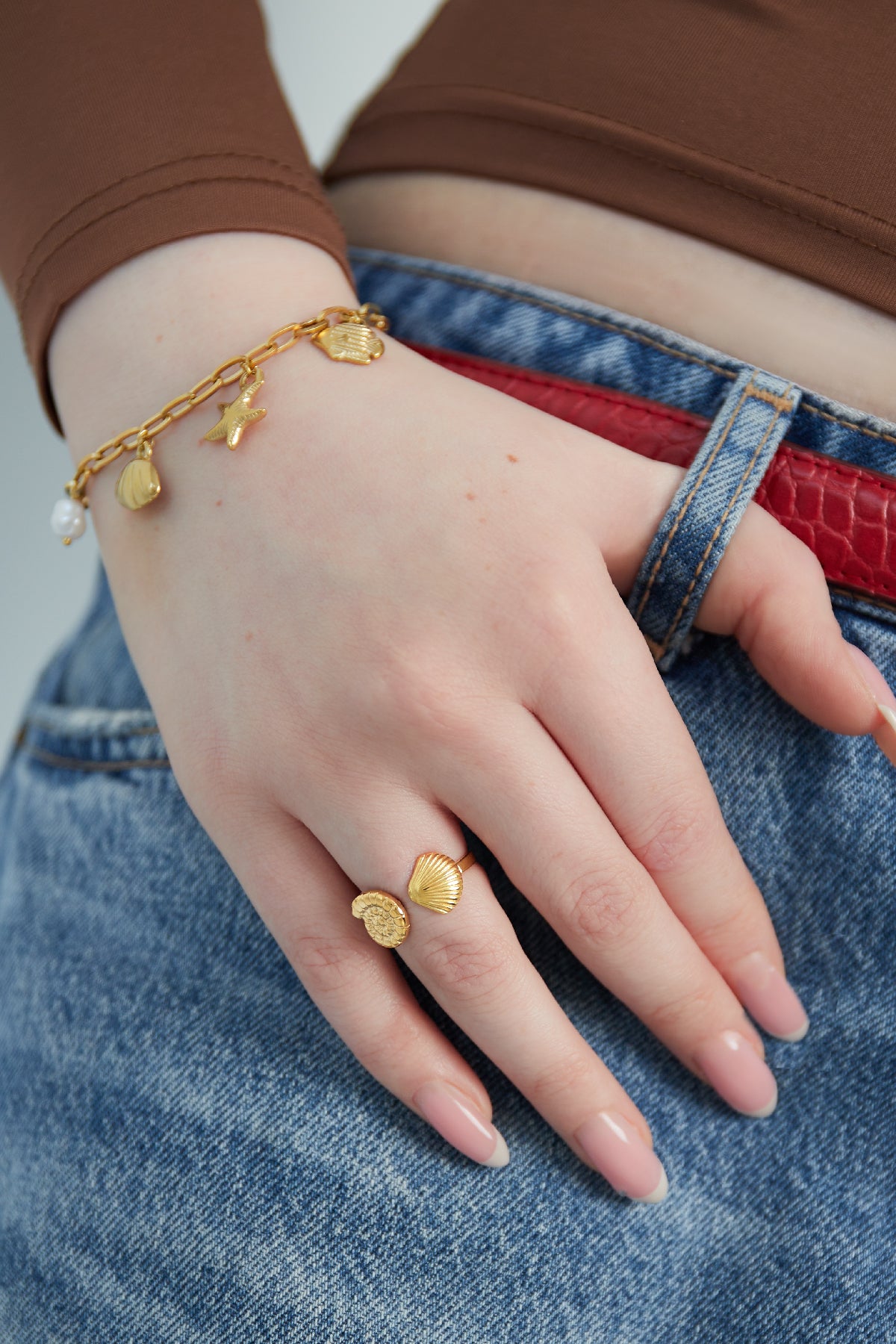 Hand model wearing gold seashell open ring paired with gold charm bracelet and brown top
