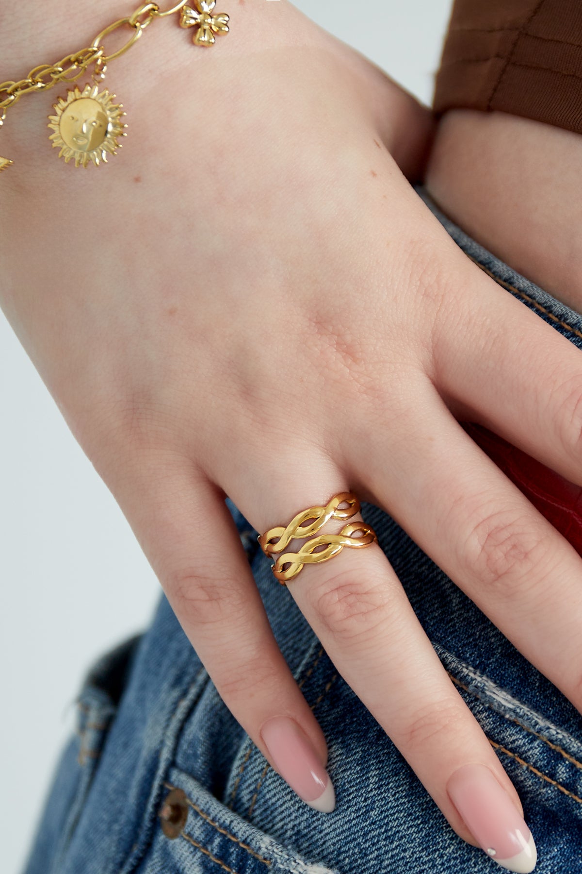 Woman's hand wearing a gold twisted double band ring paired with a sun charm bracelet and denim jeans