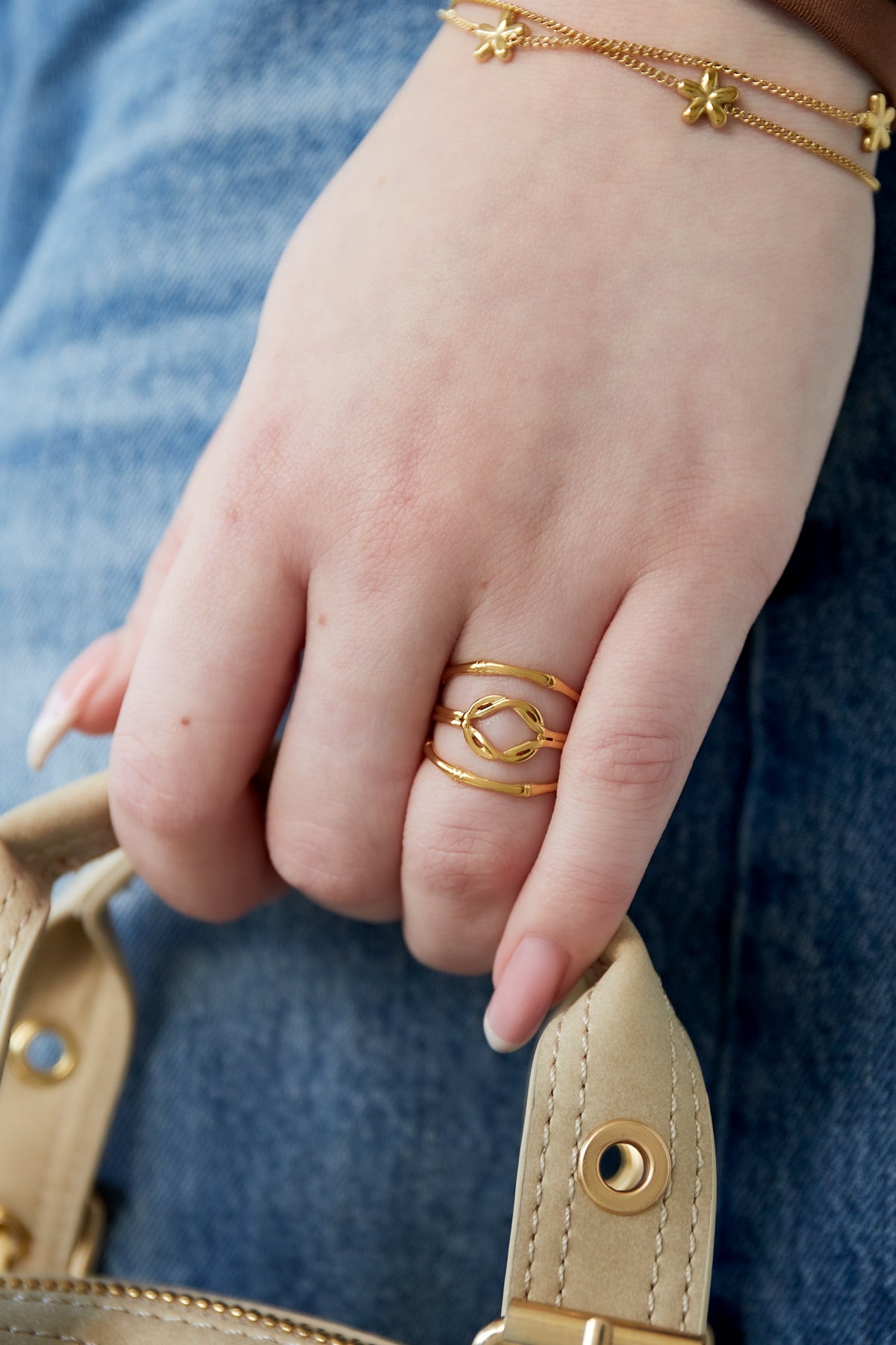 Gold layered knot ring worn on finger paired with gold flower charm bracelets, styled with casual denim and beige handbag