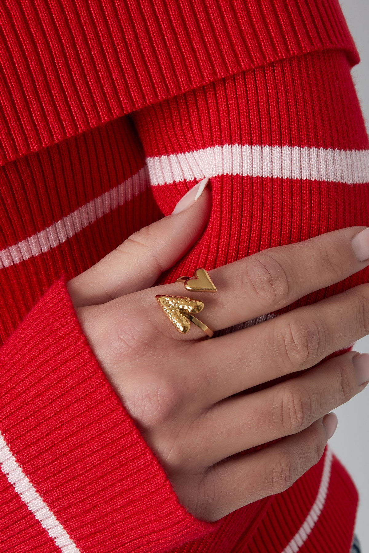 Close-up of a hand wearing the gold dual heart wrap ring paired with a red and white striped sweater