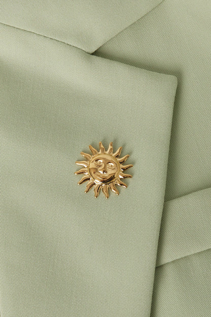Gold sun brooch pinned on the lapel of a light green blazer showing texture of the fabric and polished shine of the brooch