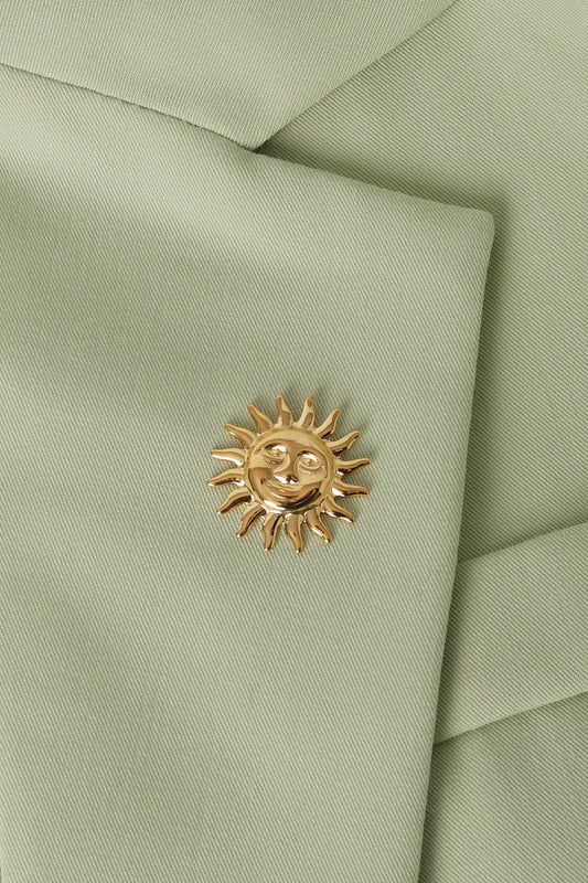 Gold sun brooch pinned on the lapel of a light green blazer showing texture of the fabric and polished shine of the brooch
