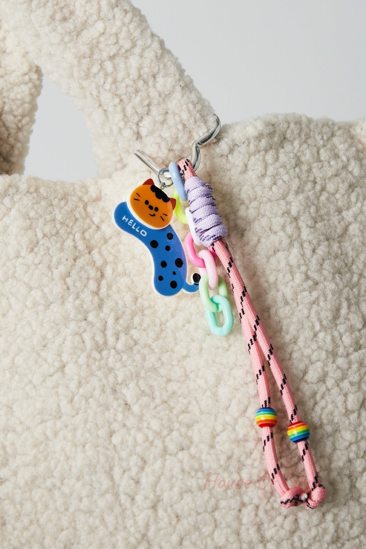 Pink cord bag charm clipped on soft cream textured fabric bag, featuring an orange cat acrylic charm with blue and pink plastic links and rainbow beads