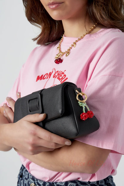 Woman holding a black textured handbag adorned with a red and green cherry bead bag charm, wearing a pink shirt