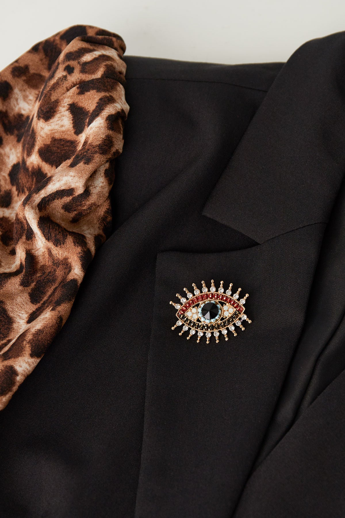 Charming eye brooch pinned on a black blazer next to a leopard print scarf, showcasing a stylish and youthful accessory look