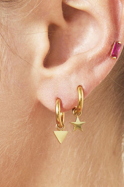Close-up of two gold hoop earrings with star and triangle charms worn on pierced ear