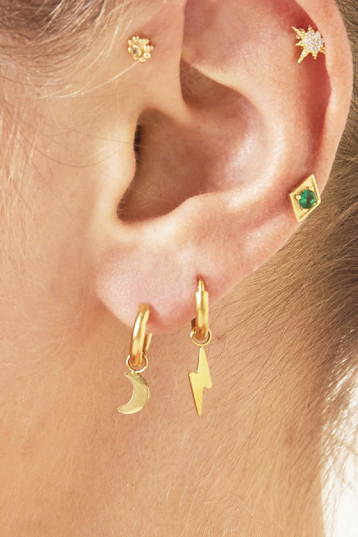 Close-up of a woman wearing mismatched gold charm hoop earrings, one with a moon and one with a lightning bolt, alongside other delicate gold studs