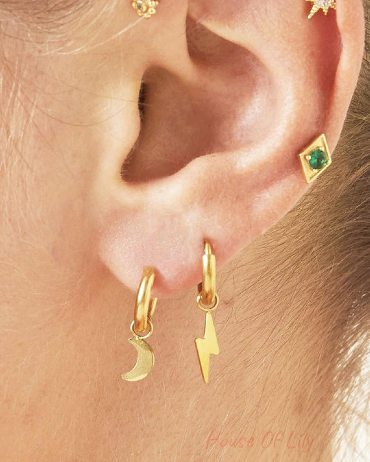 Close-up of a woman wearing mismatched gold charm hoop earrings, one with a moon and one with a lightning bolt, alongside other delicate gold studs
