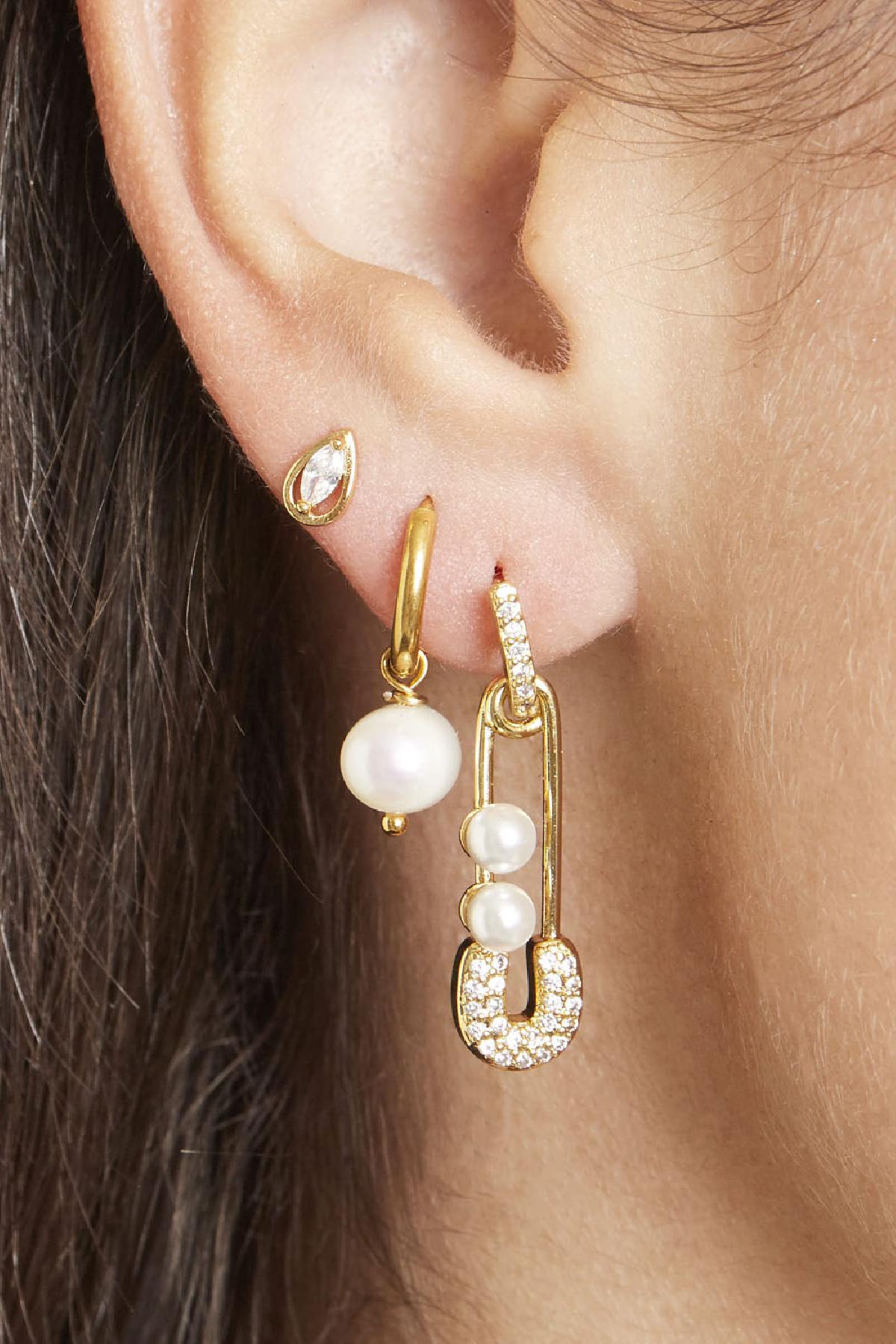 Close-up of an ear wearing three earrings: a small gold and crystal teardrop stud, a gold hoop with a single pearl charm, and a gold safety pin style earring adorned with small pearls and crystals.