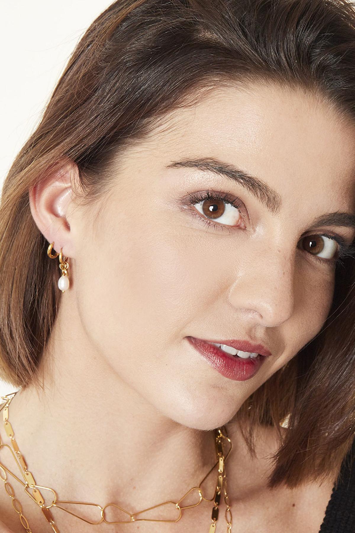 Close-up portrait of a smiling woman wearing gold pearl drop hoop earrings, styled with layered gold necklaces