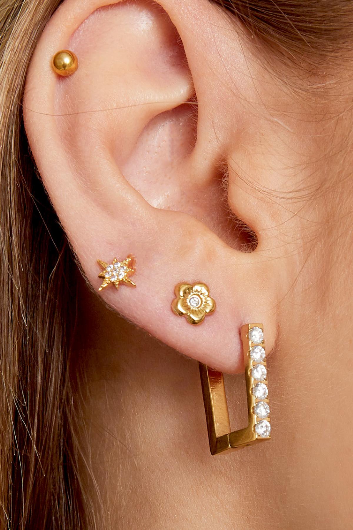 Woman's ear wearing three gold earrings including floral mini studs with rhinestone center, star-shaped and crystal-studded designs