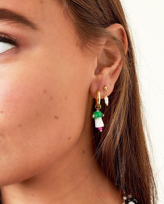 Close-up of ear with multiple earrings including gold-tone drop earring with freshwater pearl and a green mushroom charm earring