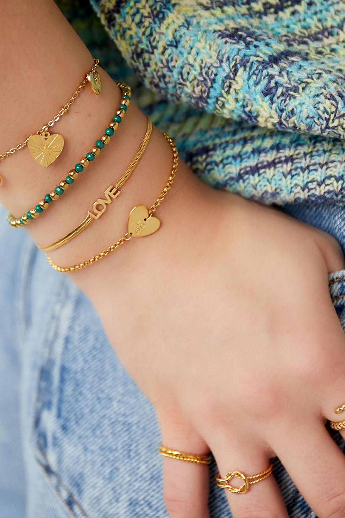 Close-up of a woman's wrist adorned with layered bracelets including the gold 'LOVE' charm bracelet, complemented by other gold and green bead bracelets, against a casual textured sweater and denim