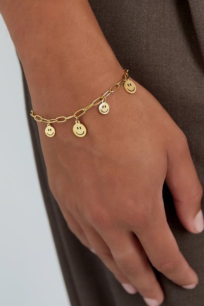 Close-up of gold smiley face charm bracelet worn on wrist with natural skin tone and neutral clothing