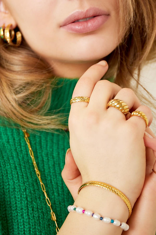 Close-up of woman wearing gold laurel cuff bracelet with rings and a pearl beaded bracelet on green sweater