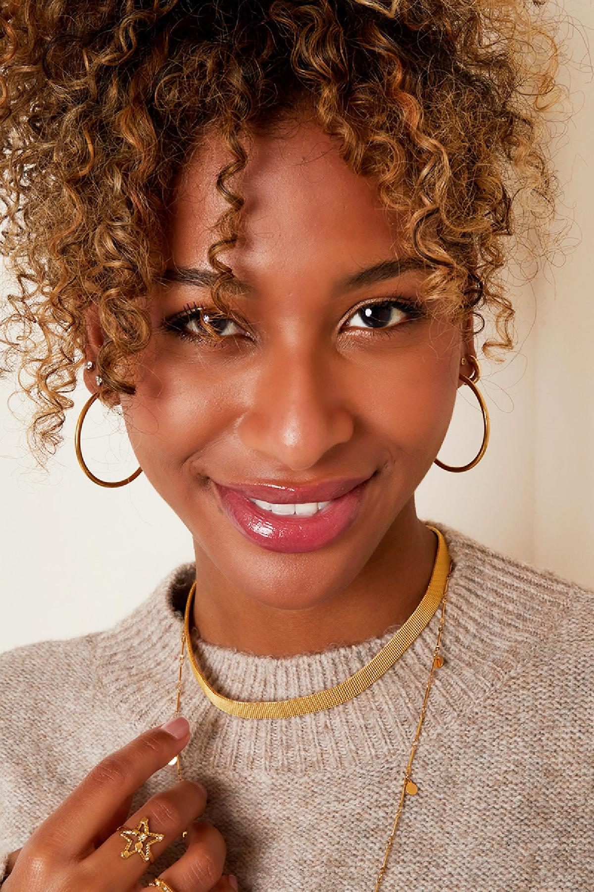 Smiling woman wearing gold hoop earrings, layered gold necklaces, beige sweater, and gold rings highlighting youthful and boutique style
