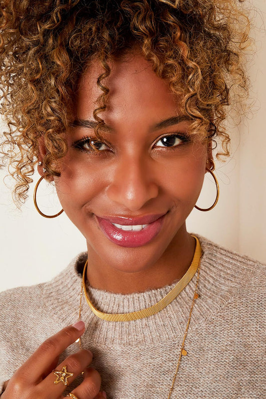 Smiling woman wearing gold hoop earrings, layered gold necklaces, beige sweater, and gold rings highlighting youthful and boutique style