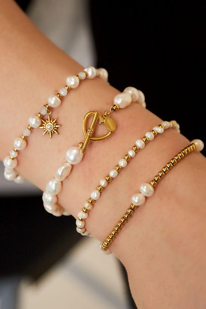 Model's wrist wearing three bracelets including the freshwater pearl bracelet with gold heart clasp, layered with complementary gold and pearl beaded bracelets.
