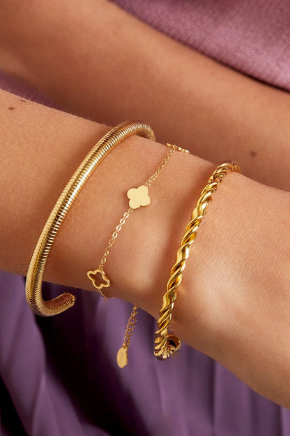 Close-up of wrist wearing gold clover charm bracelet alongside two other gold bracelets against a soft purple background