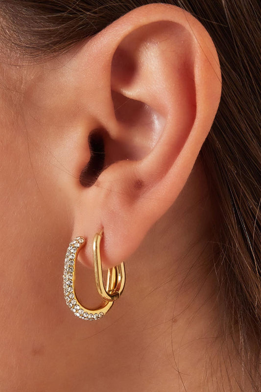 Close-up side view of a woman's ear wearing a pair of gold oval hoop earrings, one plain and one decorated with rhinestones