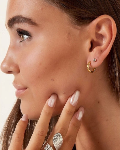Close-up of a woman wearing a gold hoop earring and a large textured silver ring on her finger, with her hand resting near her face against a soft beige background.