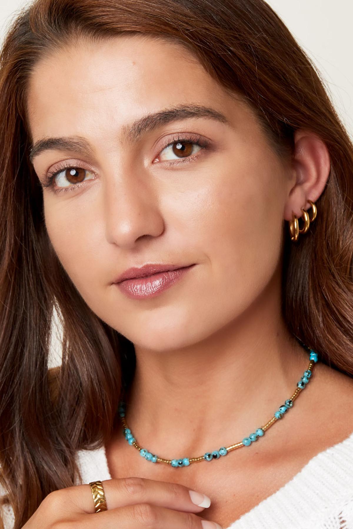 Front-facing portrait of a smiling woman wearing three gold hoop earrings and a turquoise beaded necklace