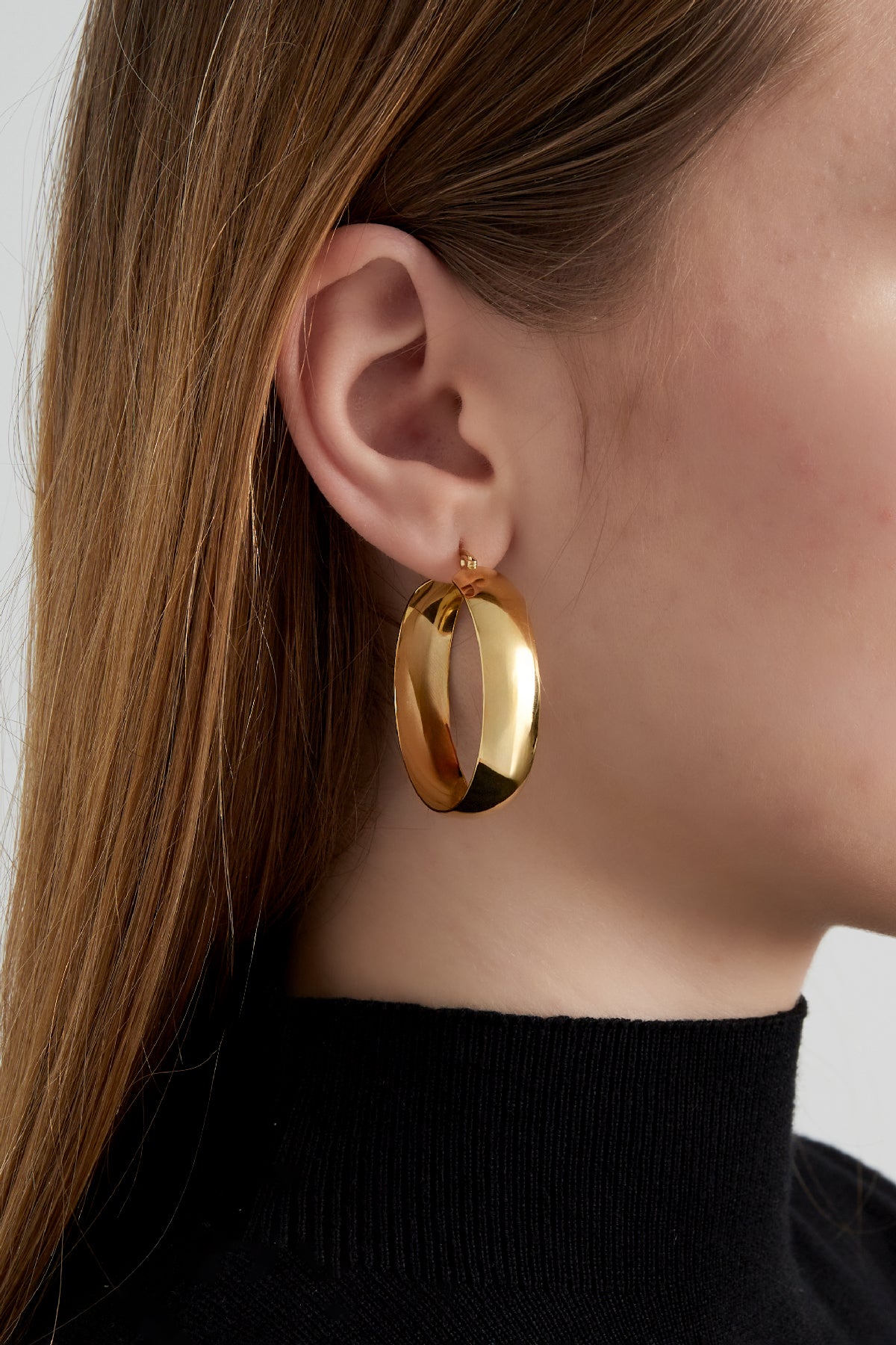 Side view of a woman wearing one wide gold hoop earring paired with a black turtleneck