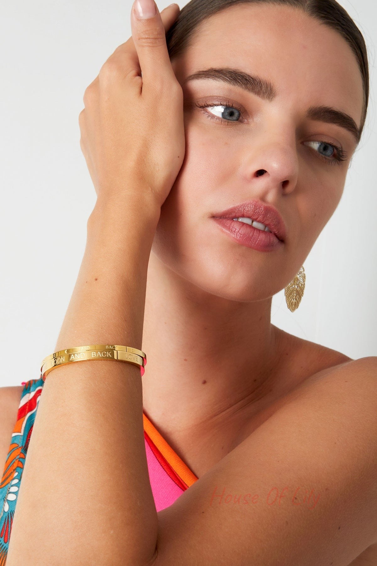 Woman wearing multiple gold engraved 'Love you to the moon and back' bracelets, styled with colorful clothing and gold leaf earrings