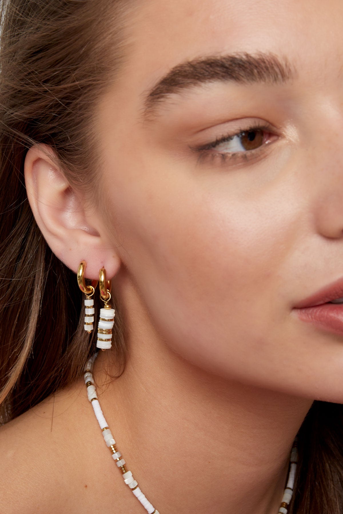 Side profile of a woman wearing two small gold hoop earrings, one with white and gold beaded pendants, paired with a matching necklace.