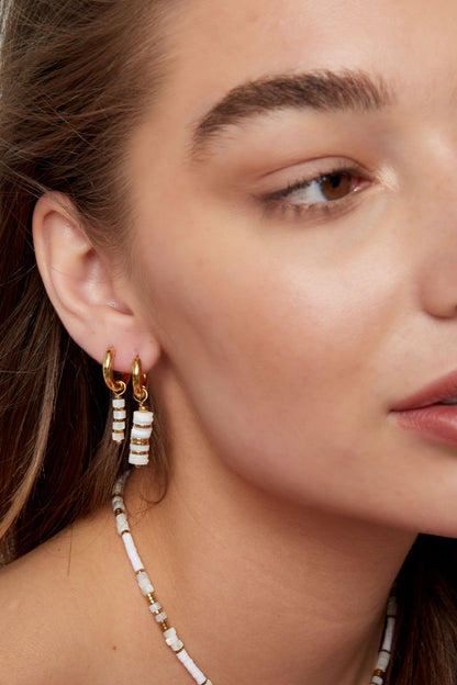 Side profile of a woman wearing two small gold hoop earrings, one with white and gold beaded pendants, paired with a matching necklace.