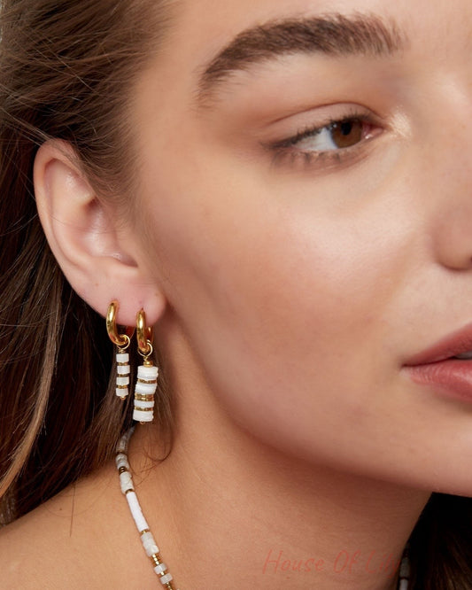 Side profile of a woman wearing two small gold hoop earrings, one with white and gold beaded pendants, paired with a matching necklace.