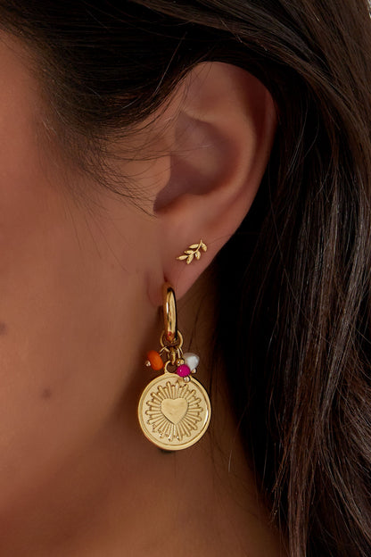 Close side view of model wearing small gold leaf stud earrings alongside statement gold charm hoops