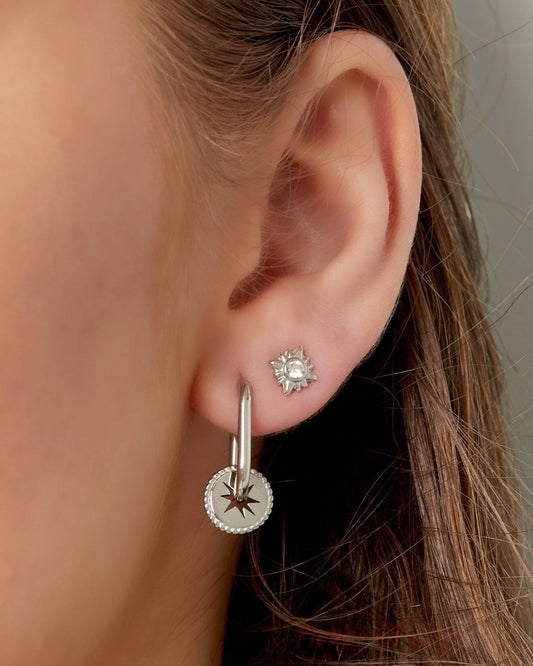 Close-up of woman wearing silver sunburst stud earrings paired with silver hoop star charm earring