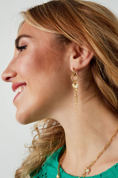 Side profile of woman wearing gold dangle hoop earrings featuring heart charm and pearl accented chains