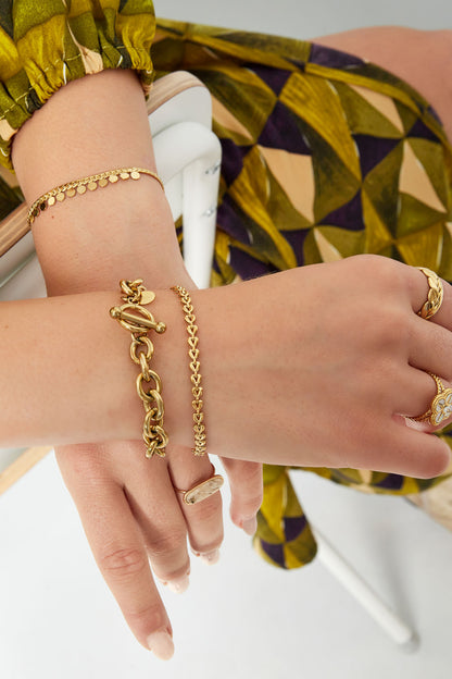 Close-up of wrist wearing two gold bracelets including linked heart chain bracelet and chunky chain bracelet against green patterned blouse