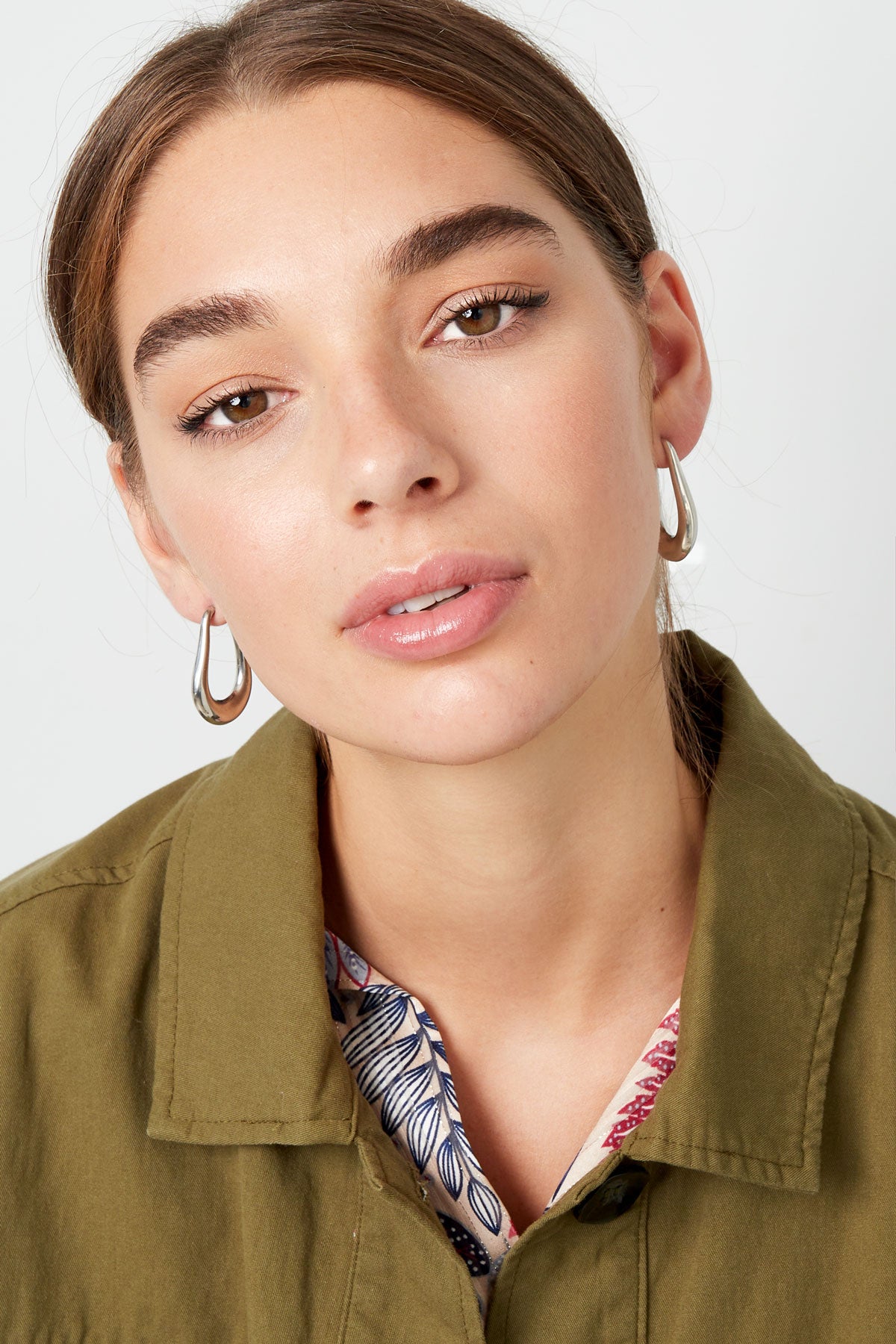 Model wearing silver teardrop hoop earrings with natural makeup and olive green jacket, front view