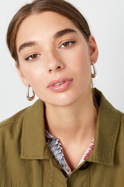 Model wearing silver teardrop hoop earrings with natural makeup and olive green jacket, front view