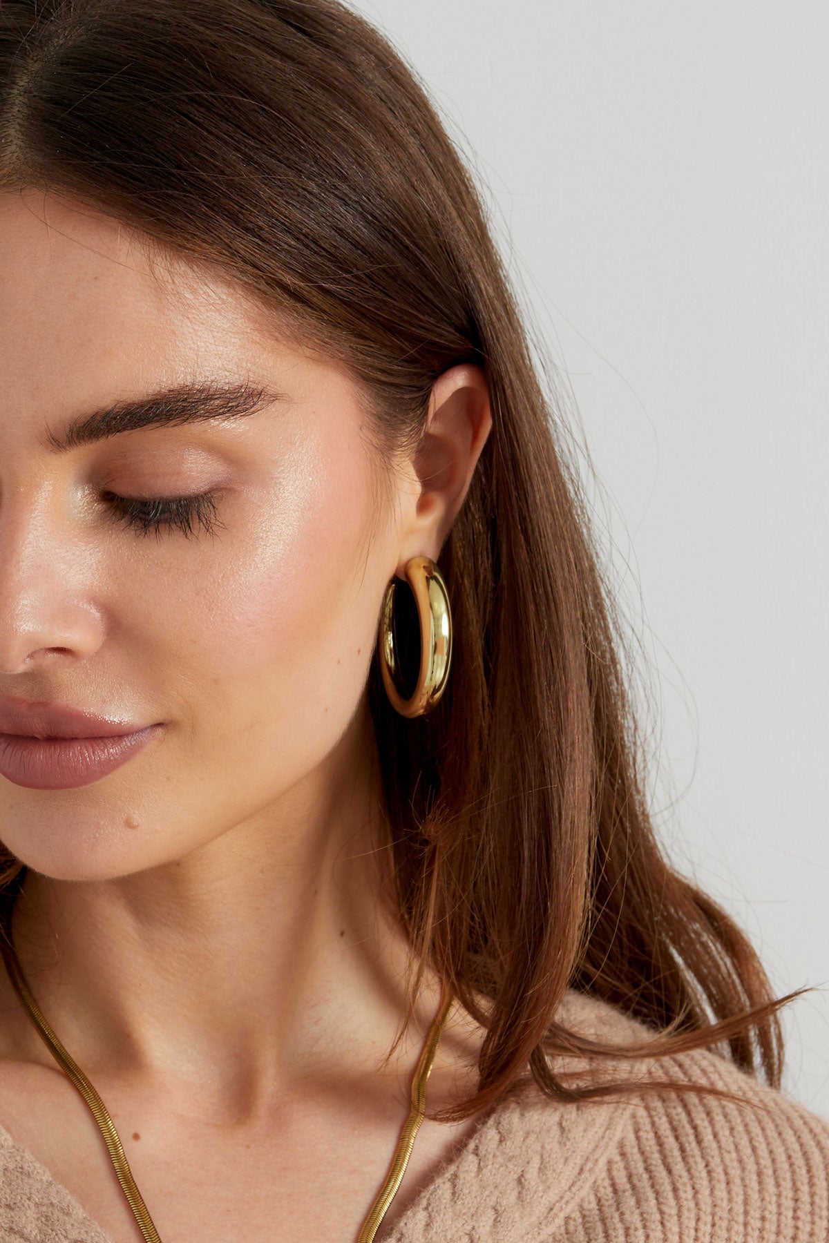 Close-up profile of woman wearing chunky gold curved hoop earrings with subtle makeup, beige knit top