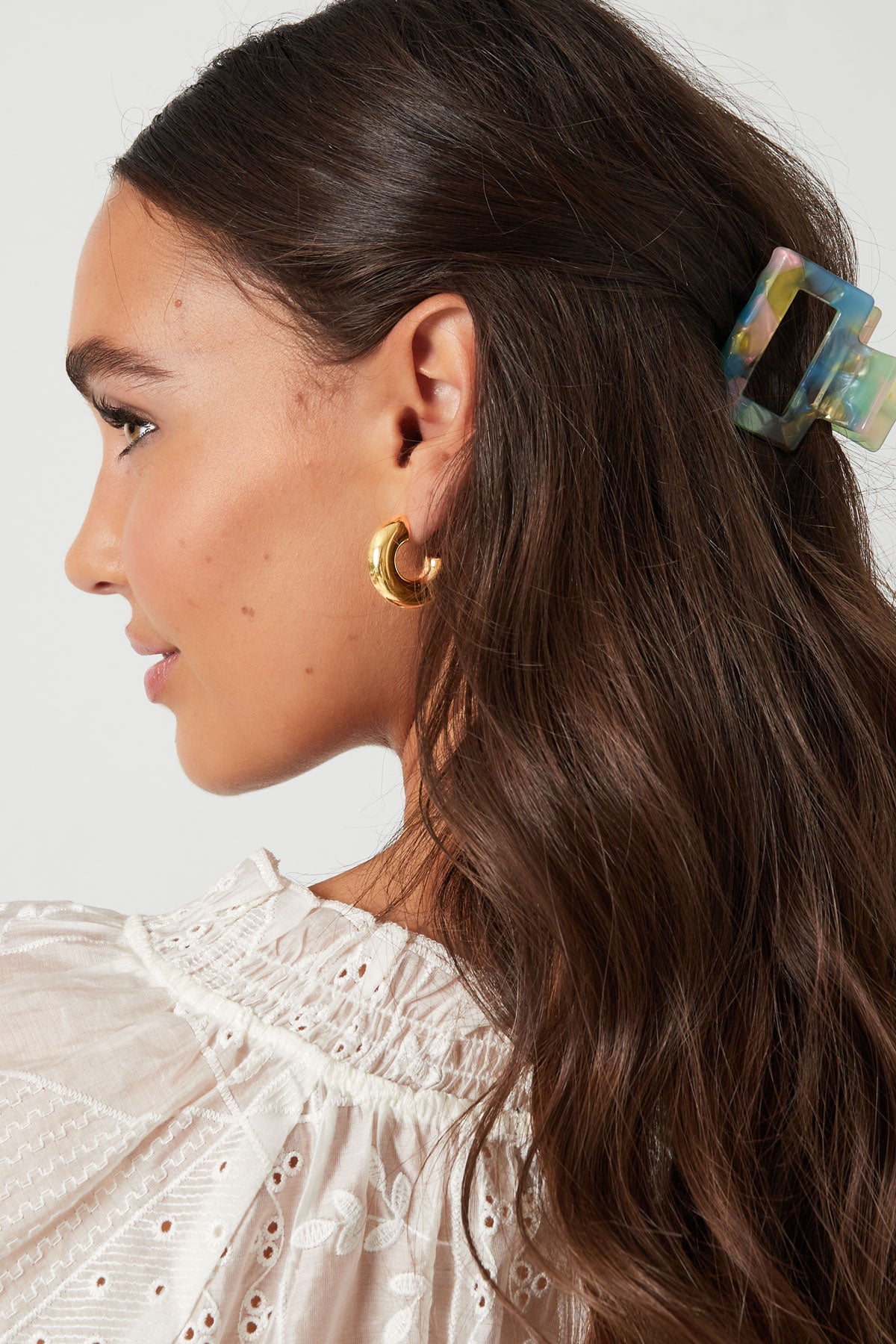 Side profile of woman wearing chunky gold hoop earrings with long wavy brown hair and white blouse