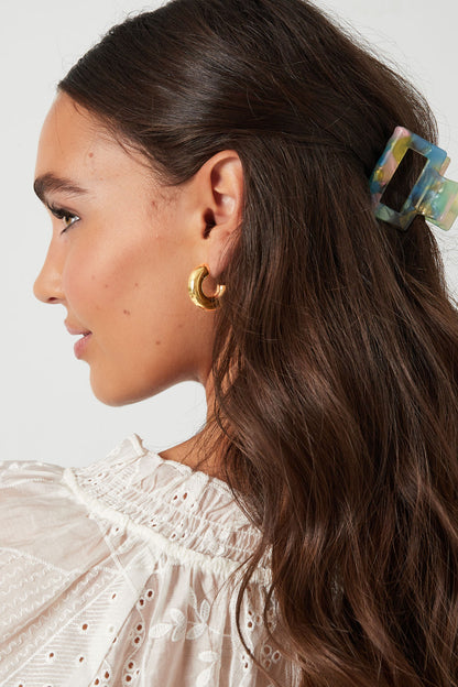 Side profile of woman wearing chunky gold hoop earrings with long wavy brown hair and white blouse