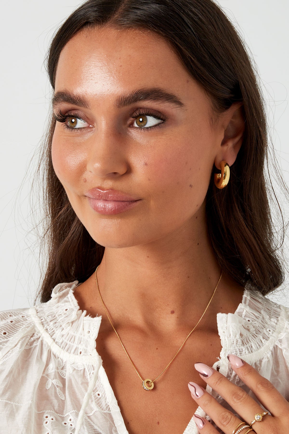 Woman facing slightly left wearing chunky gold hoop earrings and gold pendant necklace with matching rings