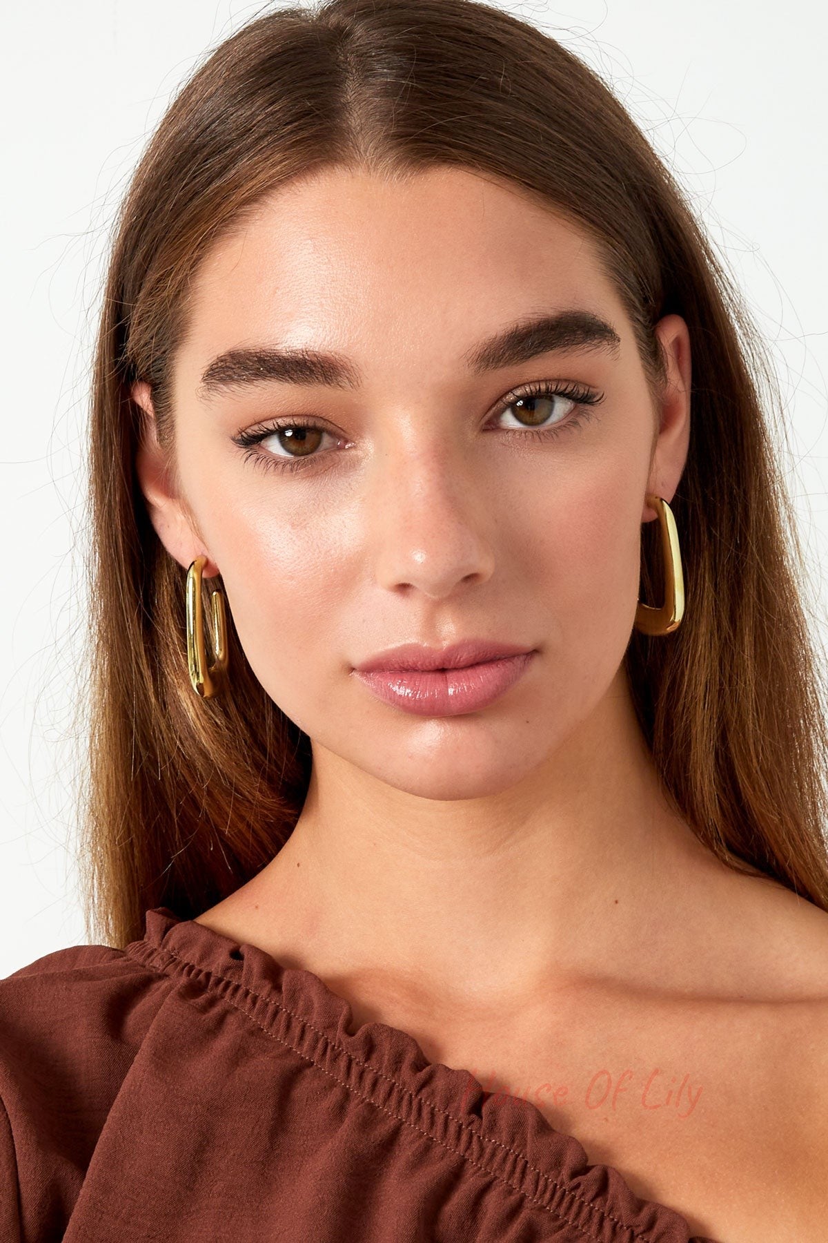 Front facing close-up of woman wearing polished gold rounded rectangle hoop earrings and brown off-shoulder top