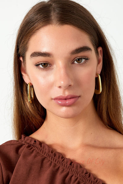 Front facing close-up of woman wearing polished gold rounded rectangle hoop earrings and brown off-shoulder top