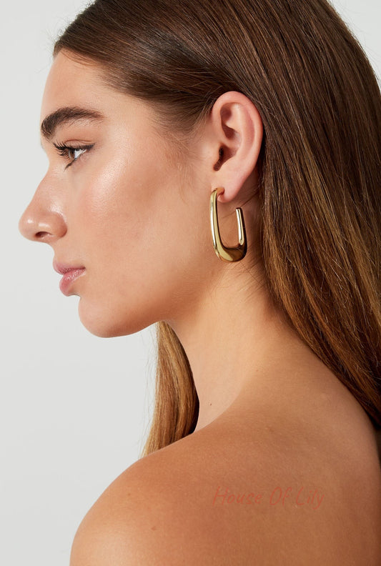 Side profile of woman wearing polished gold rounded rectangle hoop earring with natural makeup and bare shoulders