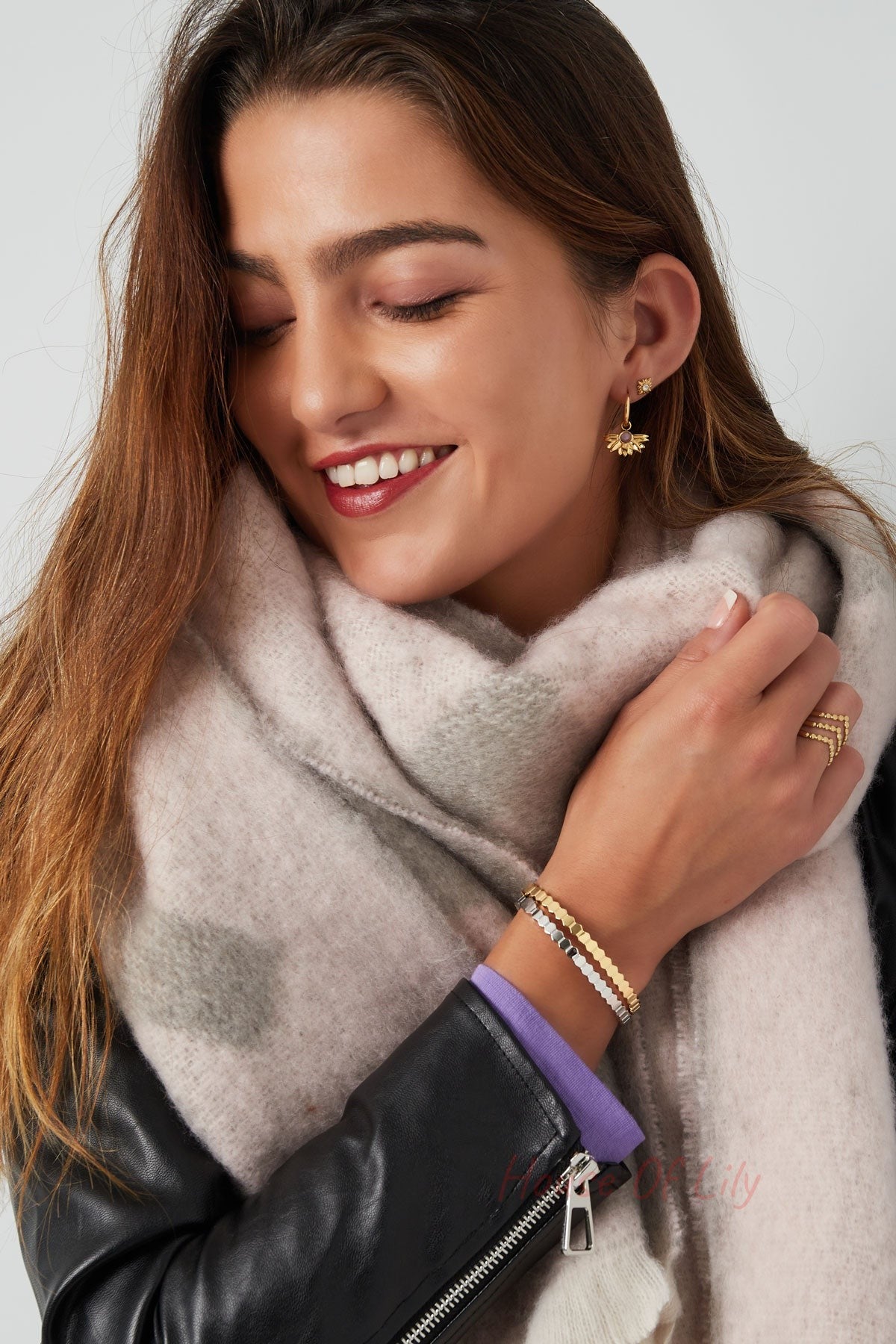 Woman wearing gold and silver hexagon link bracelets, beige scarf, black jacket smiling with eyes closed