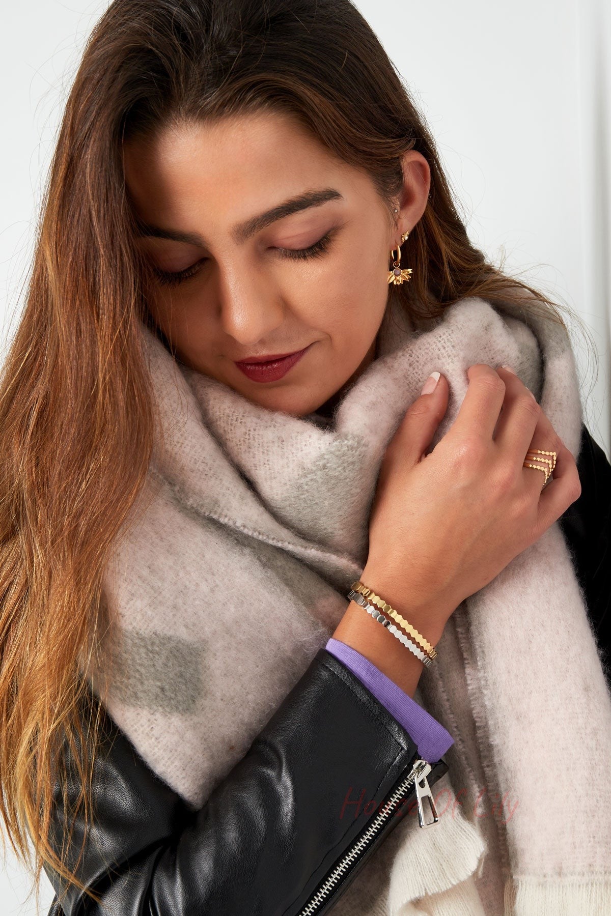 Woman showing wrist with stacked gold and silver hexagon link bracelets, beige scarf, and black jacket, head tilted down