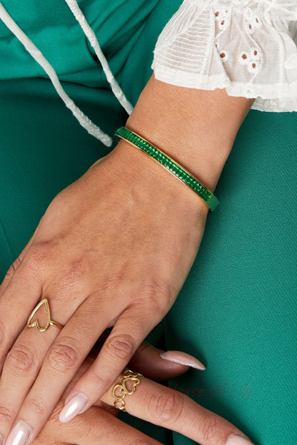 Close-up of wrist wearing a gold-tone bangle bracelet with green square-cut crystals paired with gold heart rings and teal outfit