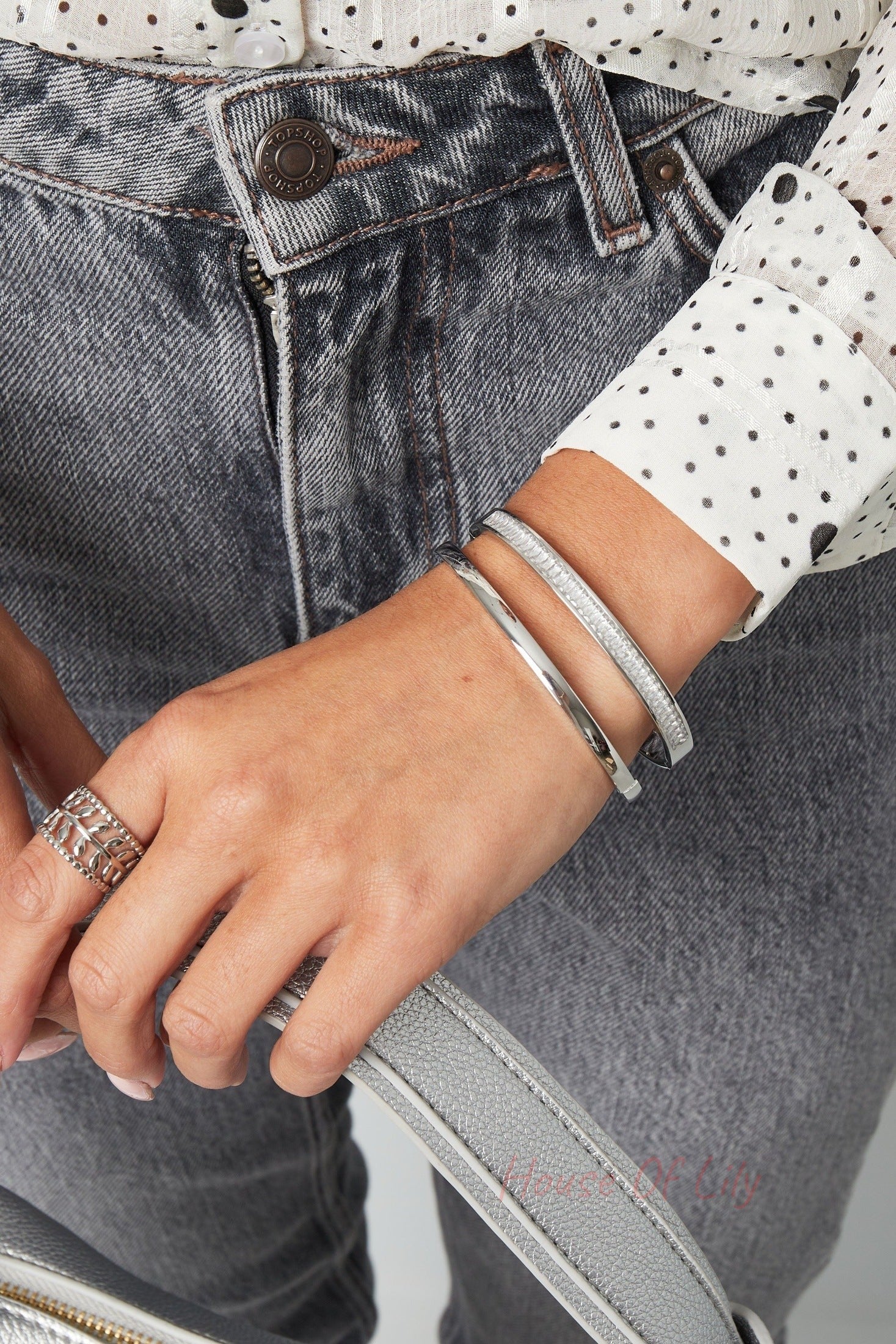 Woman's wrist wearing stacked slim silver bracelets paired with silver ring and gray jeans